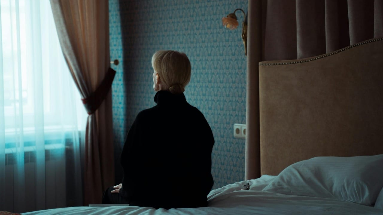 A person with blonde hair in a black sweater sits on a bed facing a window.