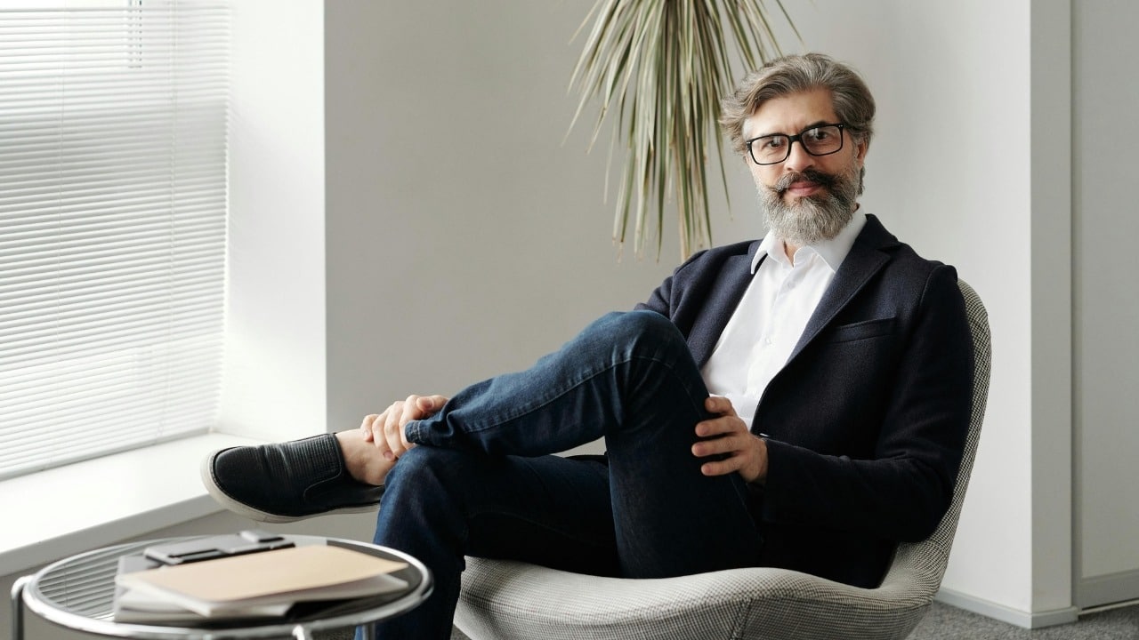 A man with a grey beard and glasses sits in a chair with his legs crossed.