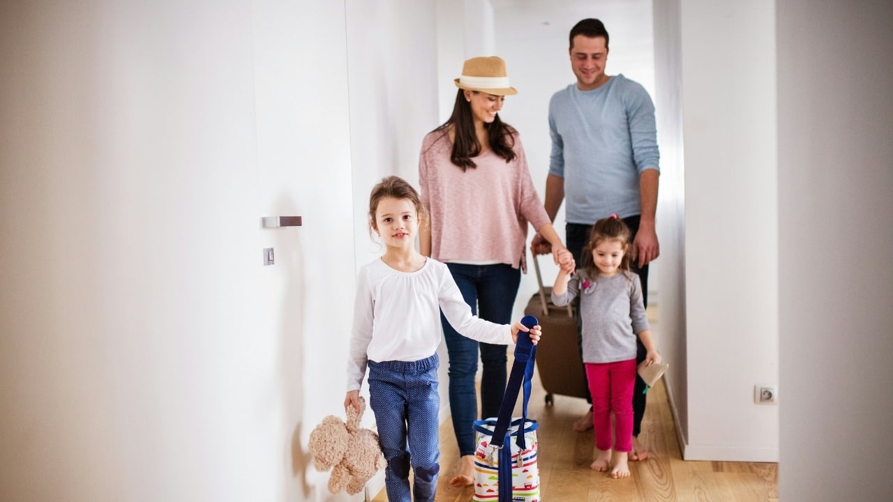 A family of four walks down a brightly lit hallway while carrying luggage and a teddy bear.