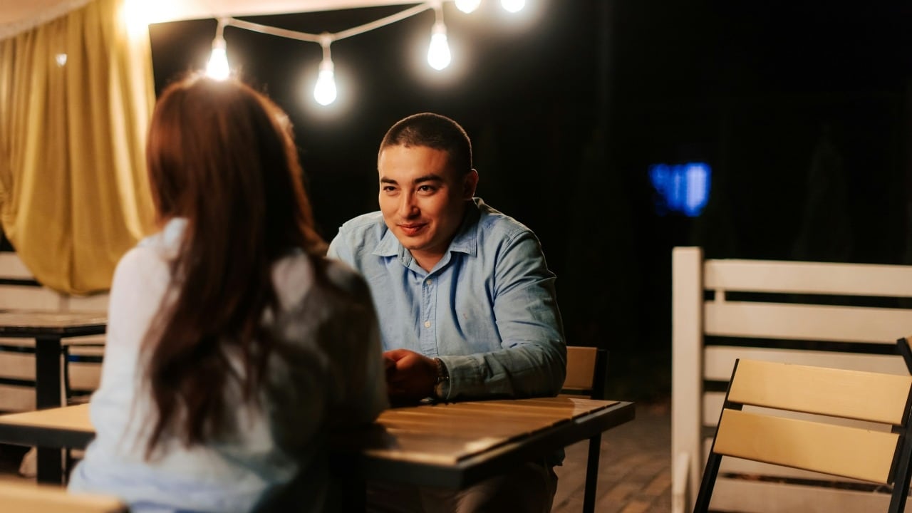 A man in a blue shirt smiles while sitting at an outdoor table across from a woman.