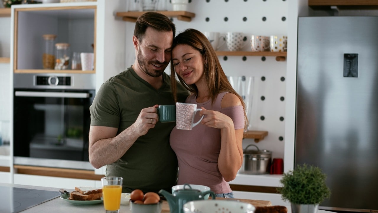A smiling couple stands in a kitchen together while holding mugs of coffee.