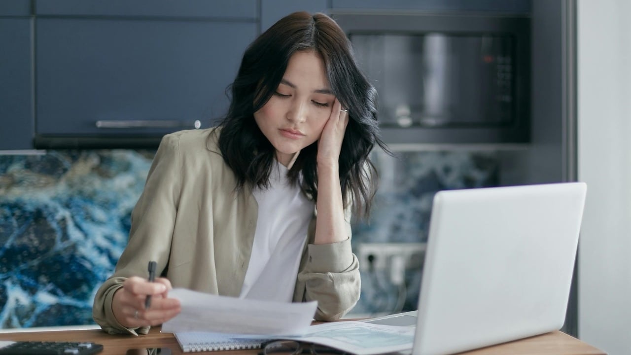 A woman with dark hair sits at a desk looking stressed while reviewing paperwork.