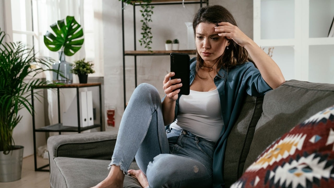 A woman sits on a grey couch looking down at her smartphone with a concerned expression.