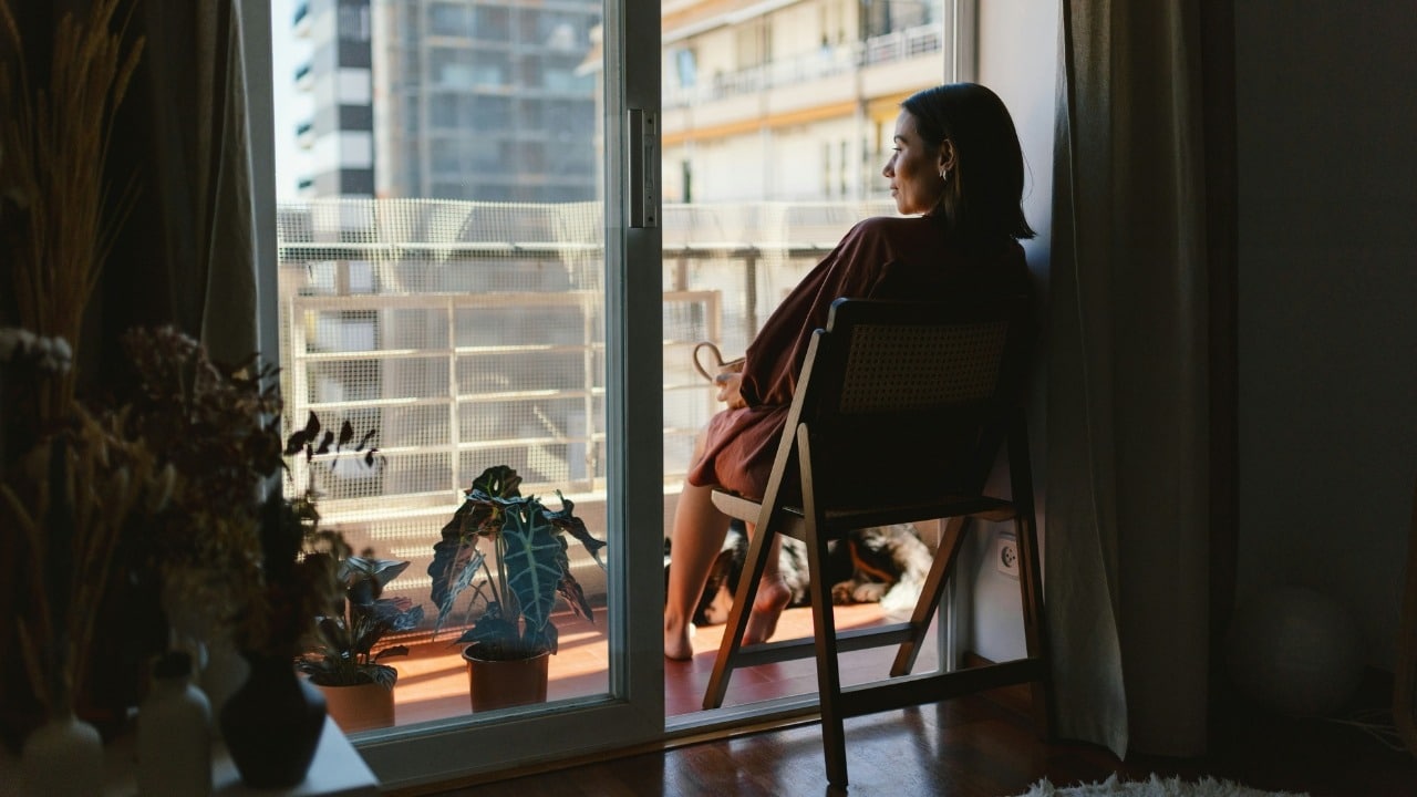 A woman sits on a wooden chair on a balcony overlooking tall buildings in the city distance.