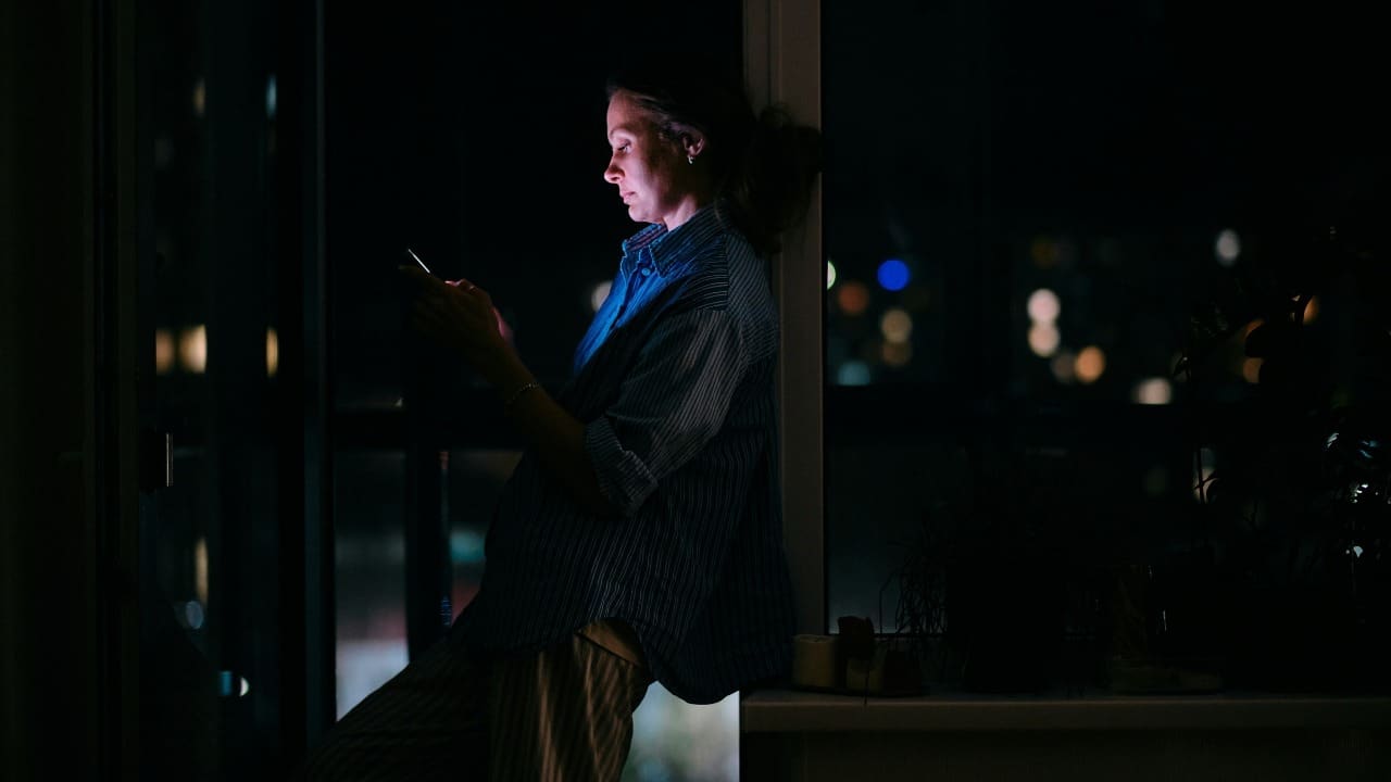 A woman leans against a window frame at night while looking at her glowing smartphone screen.