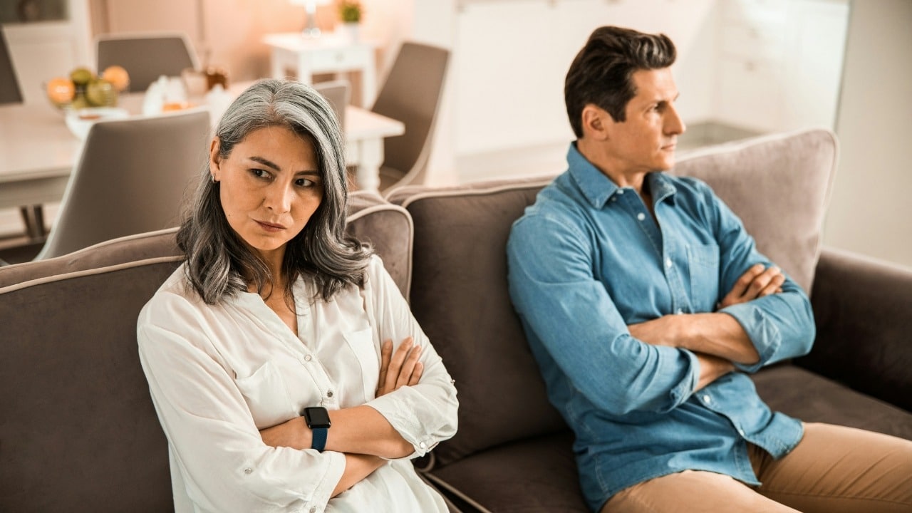A man and woman sit on a sofa with arms crossed, looking away from each other.