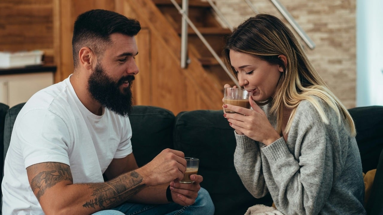 A bearded man watches a woman drink from a small glass while sitting on a sofa.