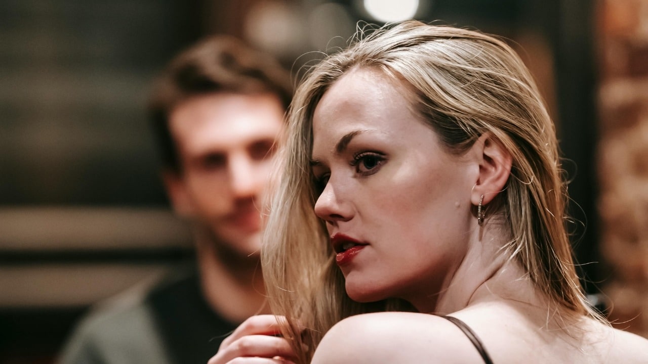 A woman with blonde hair looks over her shoulder while a man stands blurred behind her.