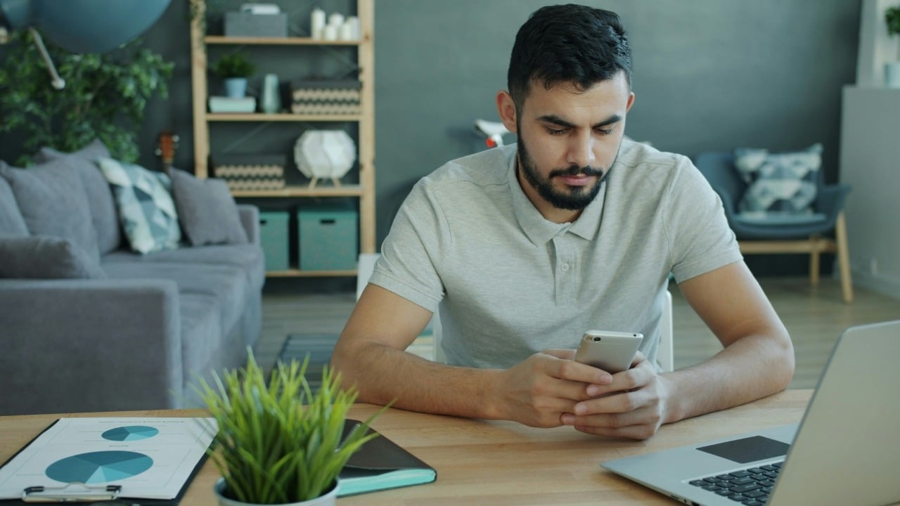 A man sits at a desk with a laptop and documents while holding a smartphone.