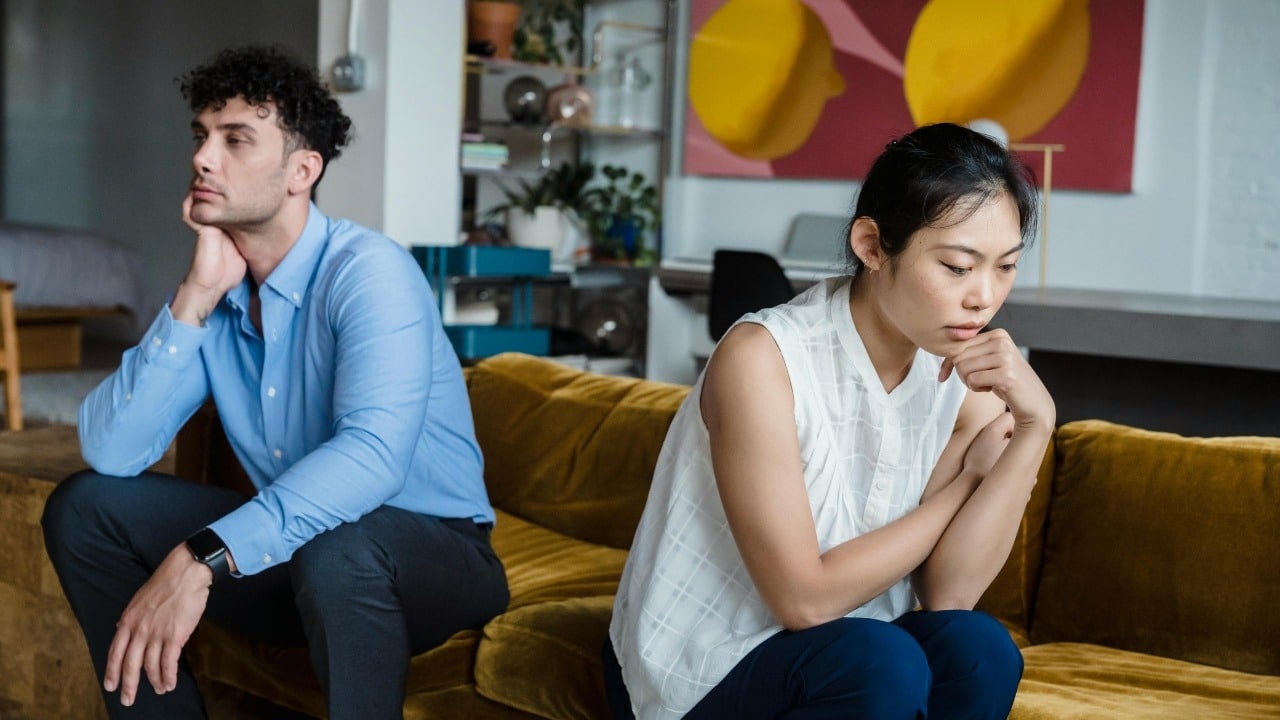 A woman sits on a bed looking away while a man sits behind her looking down.