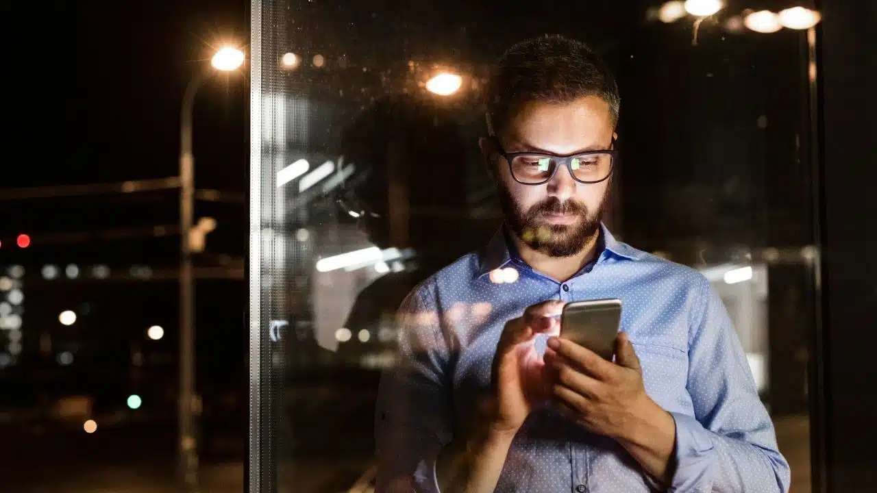A man with glasses uses his phone at night, standing next to a glass window.