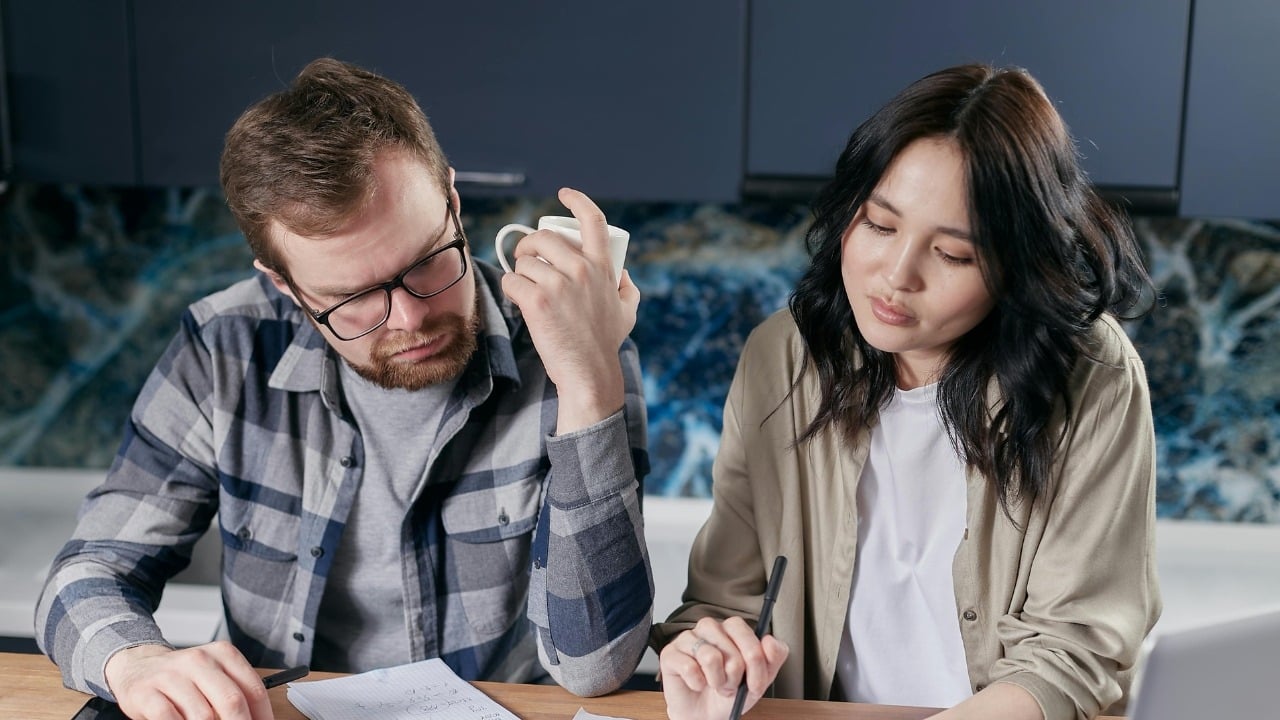 A man and woman sit at a table looking down at papers while the man holds a mug.