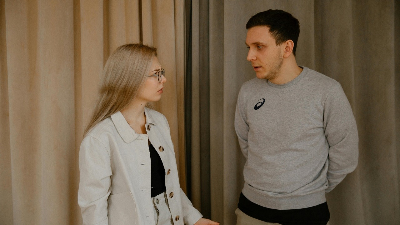 A woman wearing glasses looks at a man in a grey sweatshirt standing in front of curtains.