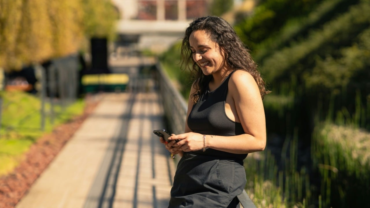 A smiling woman with curly hair stands on a walkway and looks at her phone.