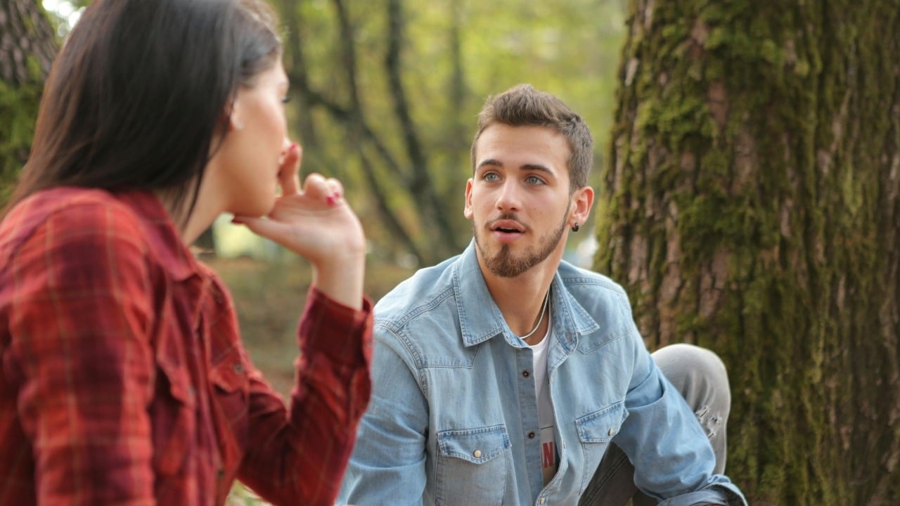 A man in a denim shirt looks at a woman sitting near a mossy tree.