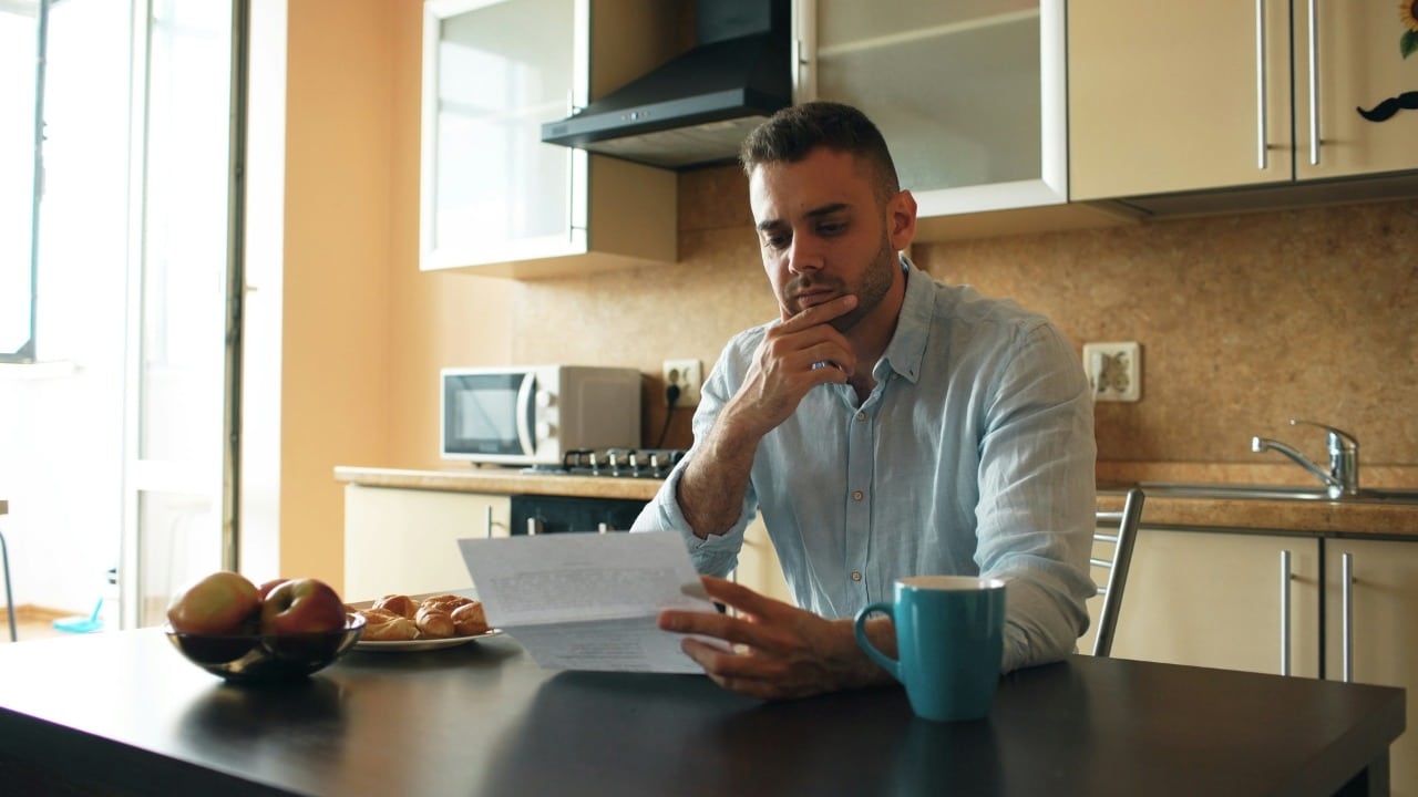 A man in a blue shirt sits in a kitchen reading a paper with a pensive expression.