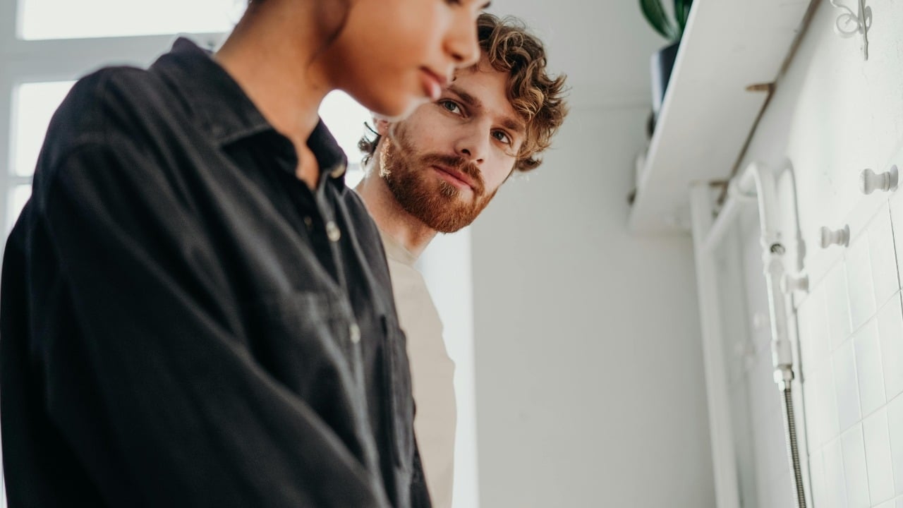 A man with a beard looks toward a woman in a black shirt in a kitchen.