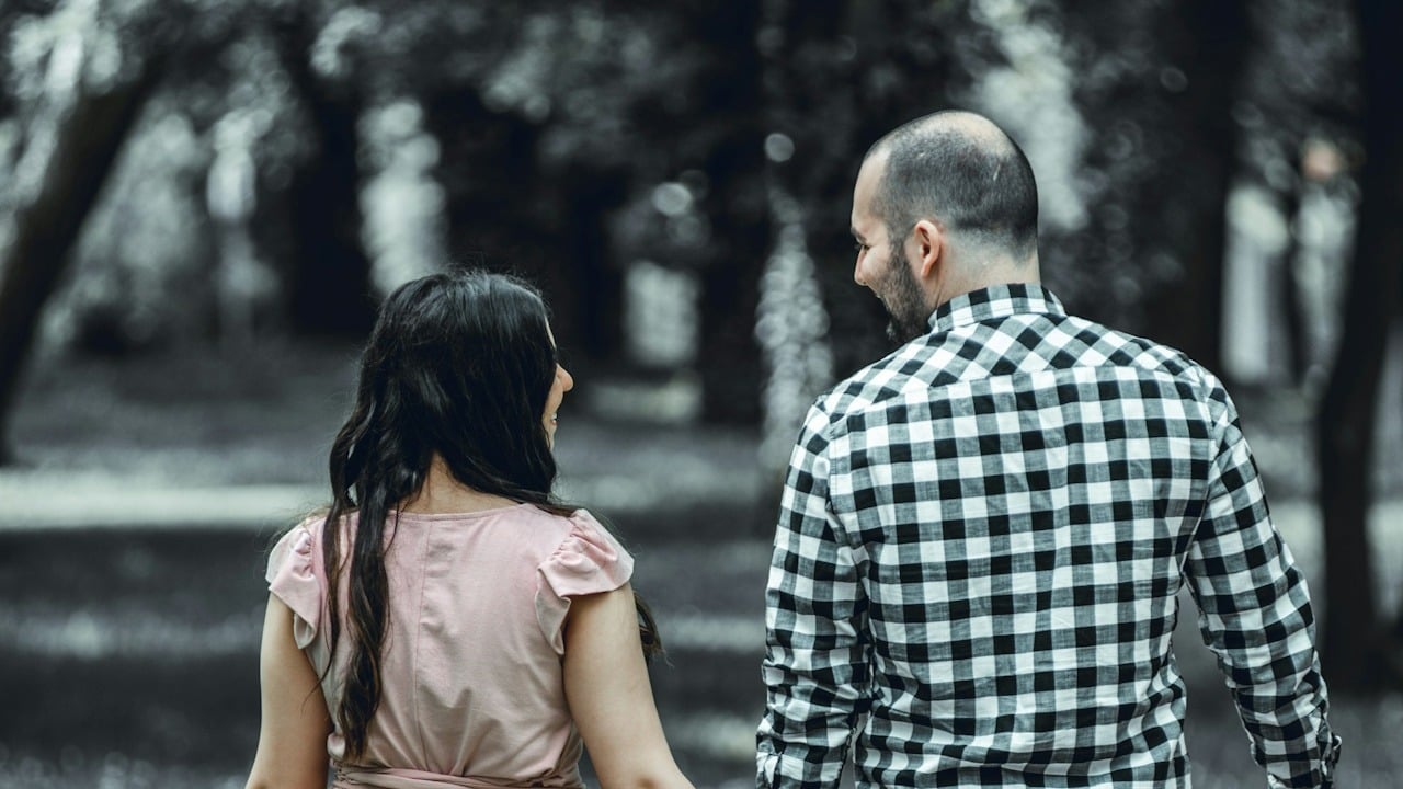 A balding mature man with a light beard walking down a path with a younger woman as they smile at each other and have their backs to the camera.