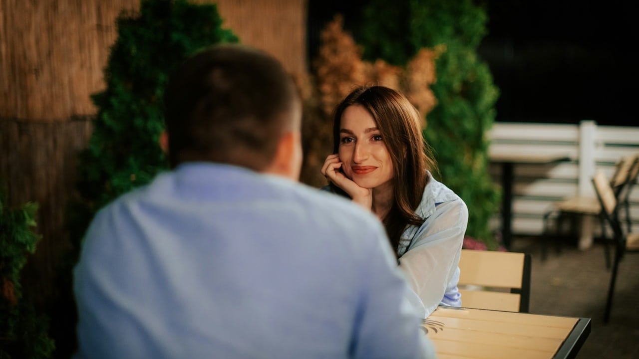 A smiling woman staring at a man and holding her hand in her palm as they are seated at a table.