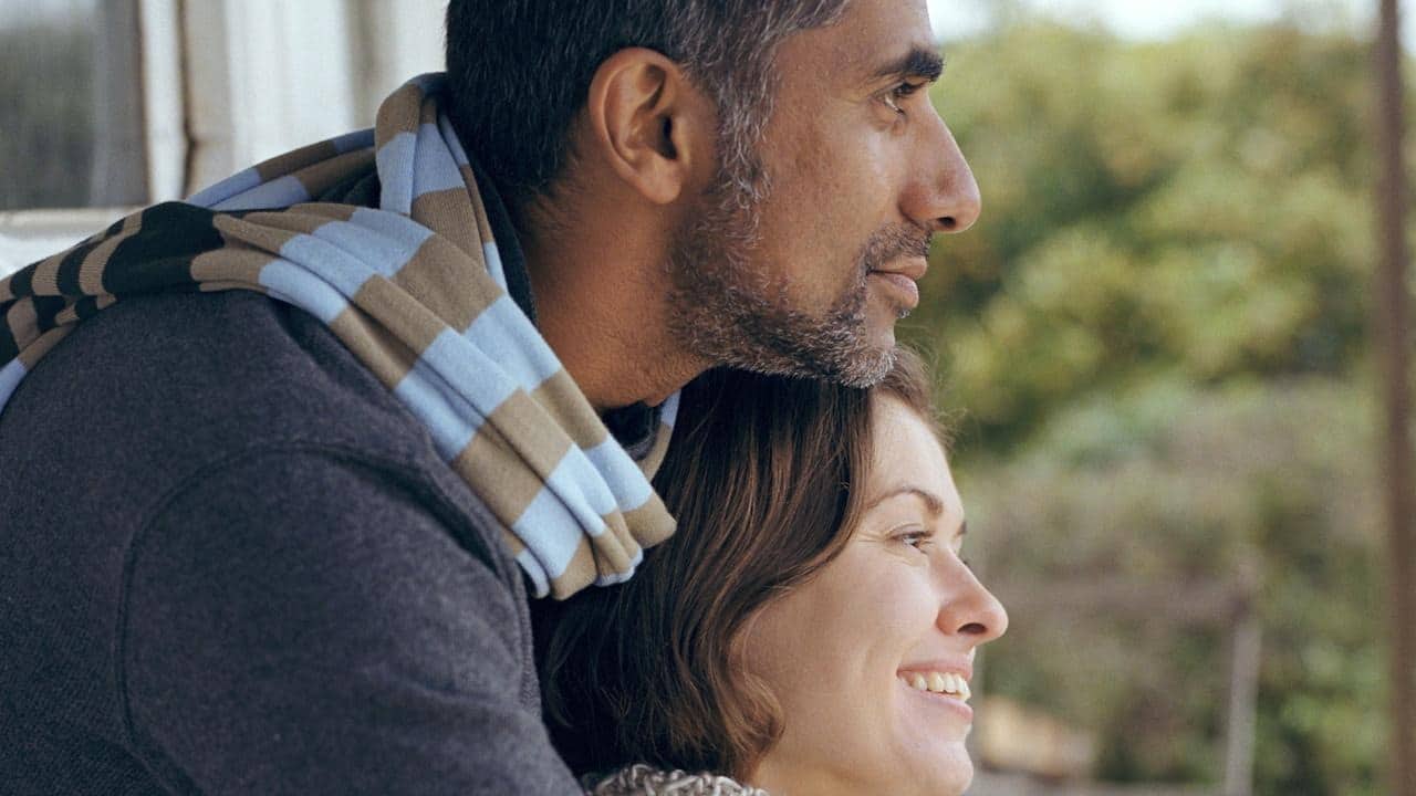 A woman resting her head against a man with a greying beard’s chest as they gaze outwards.