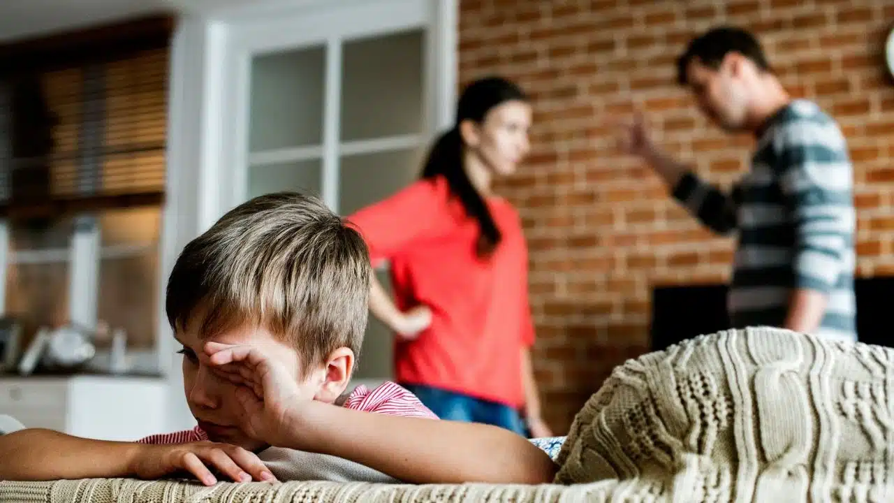 A young boy wipes his eyes while two adults stand blurred in the background.