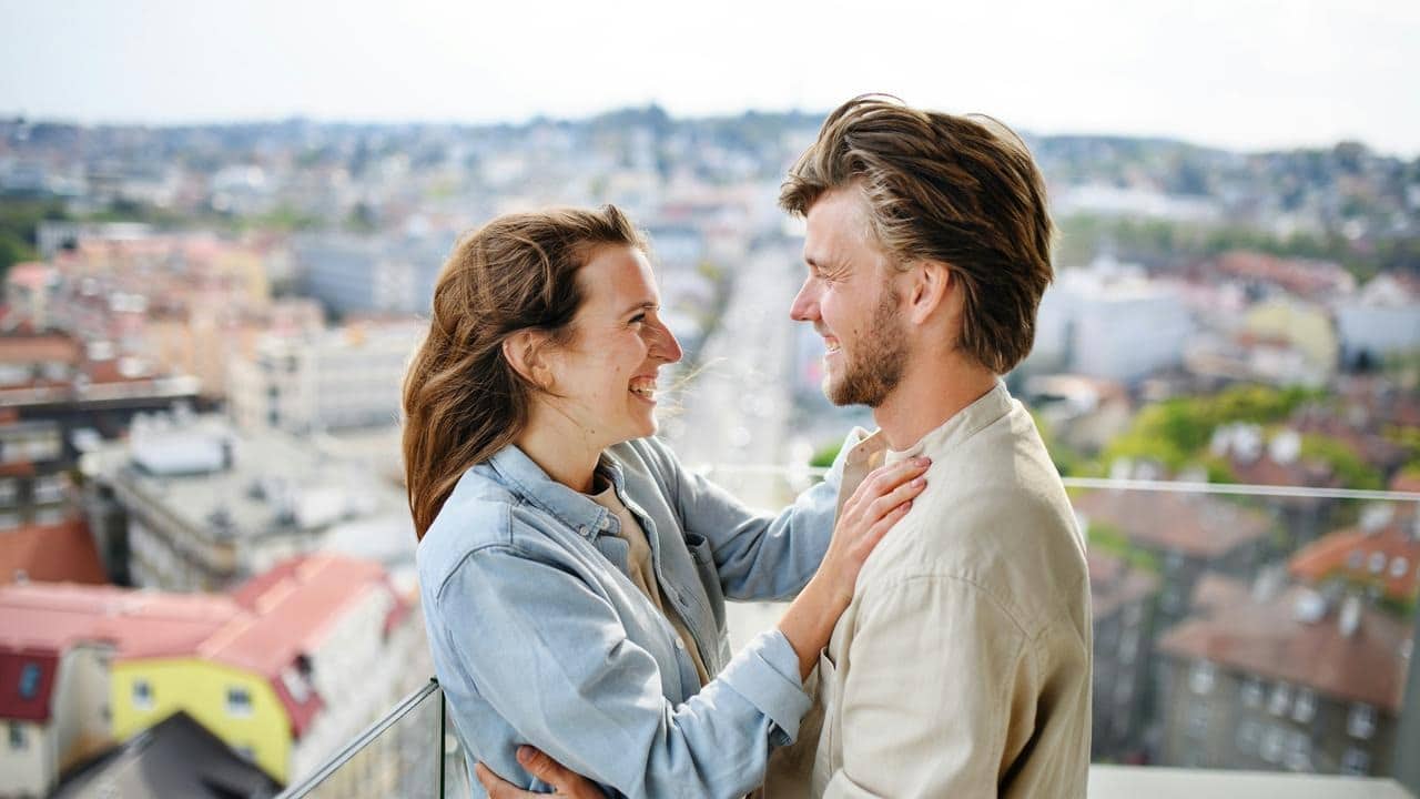 A smiling couple standing close together on a balcony overlooking a city.