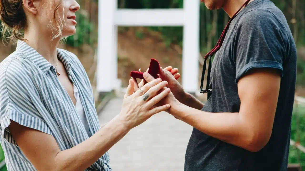 Two people hold a small red box containing a ring while standing outdoors on a bridge.