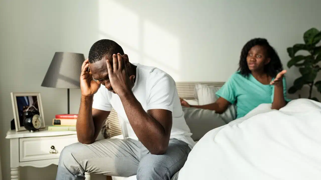 A man sits on a bed with hands on his head while a woman gestures behind him.