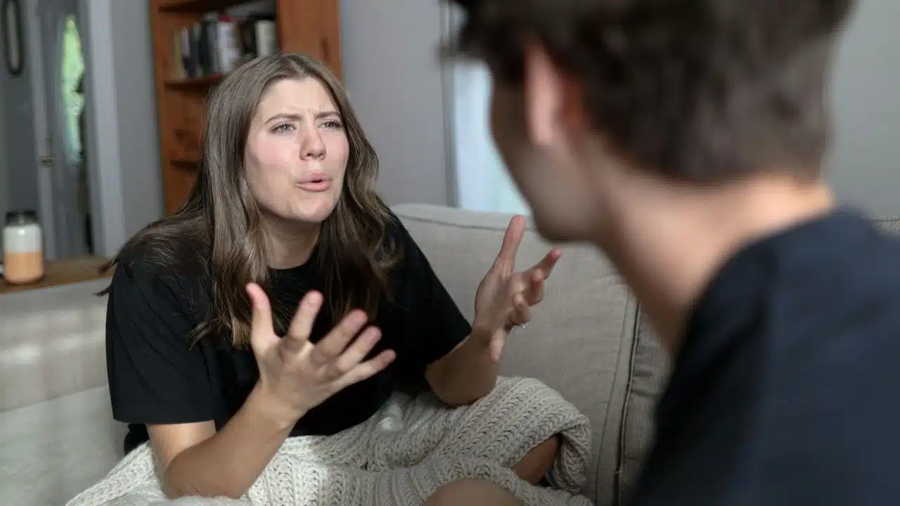 A woman sitting on a couch with a blanket gestures emphatically while speaking to a man.