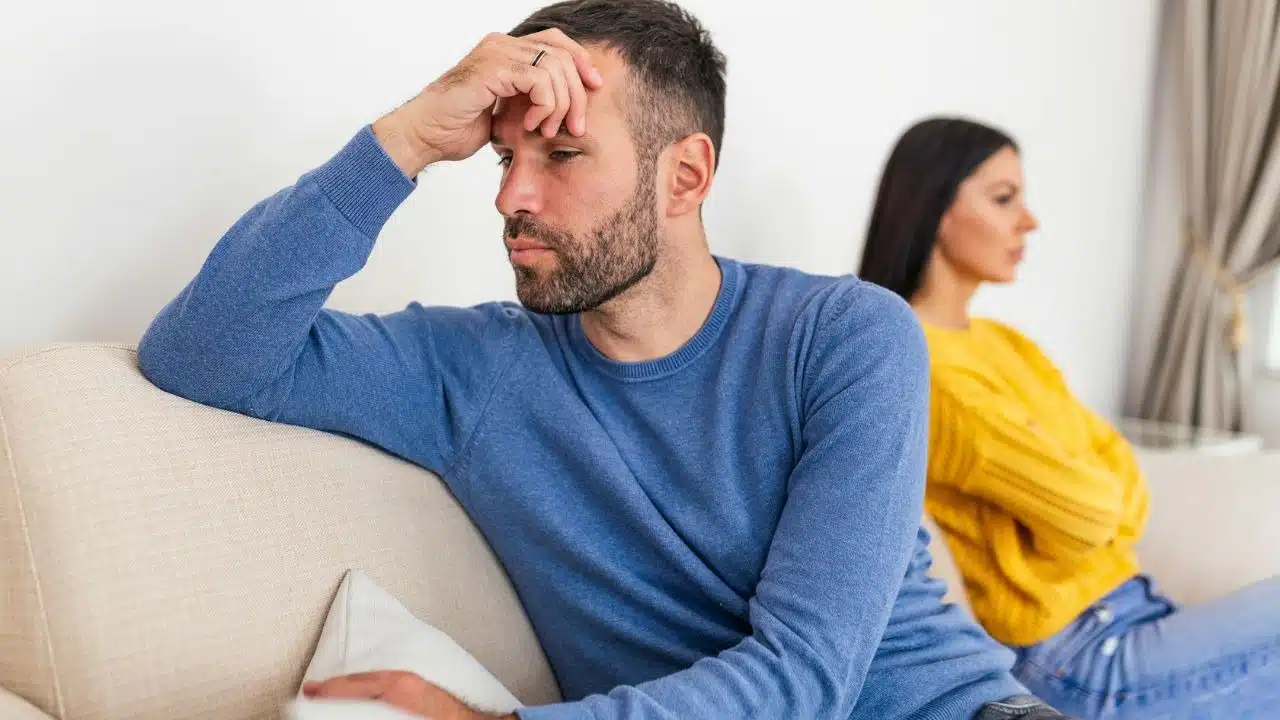 A man in a blue sweater sits on a sofa looking distressed while a woman sits behind him.