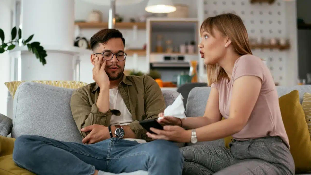 A woman gestures with a tissue while speaking to a man sitting on a grey sofa.