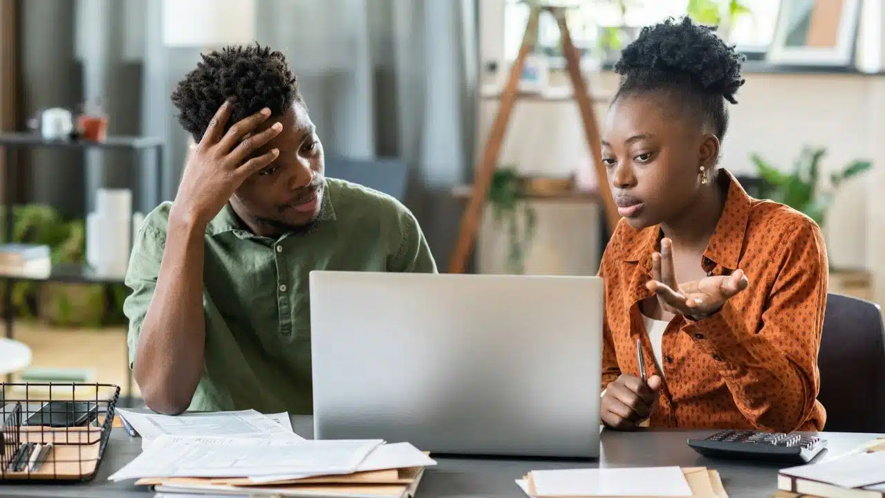A man looks stressed while a woman gestures toward a laptop and several financial documents.