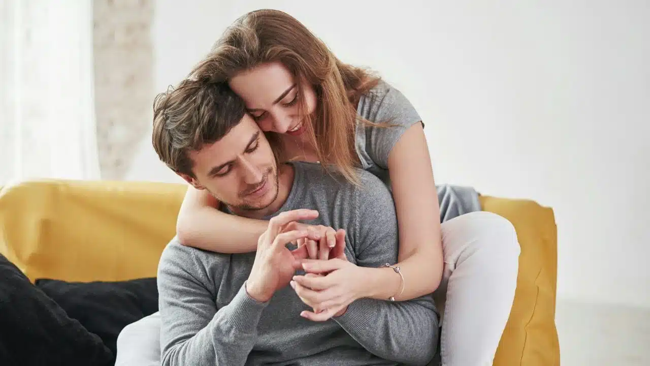 A woman hugs a man from behind while they both look at a ring on his finger.