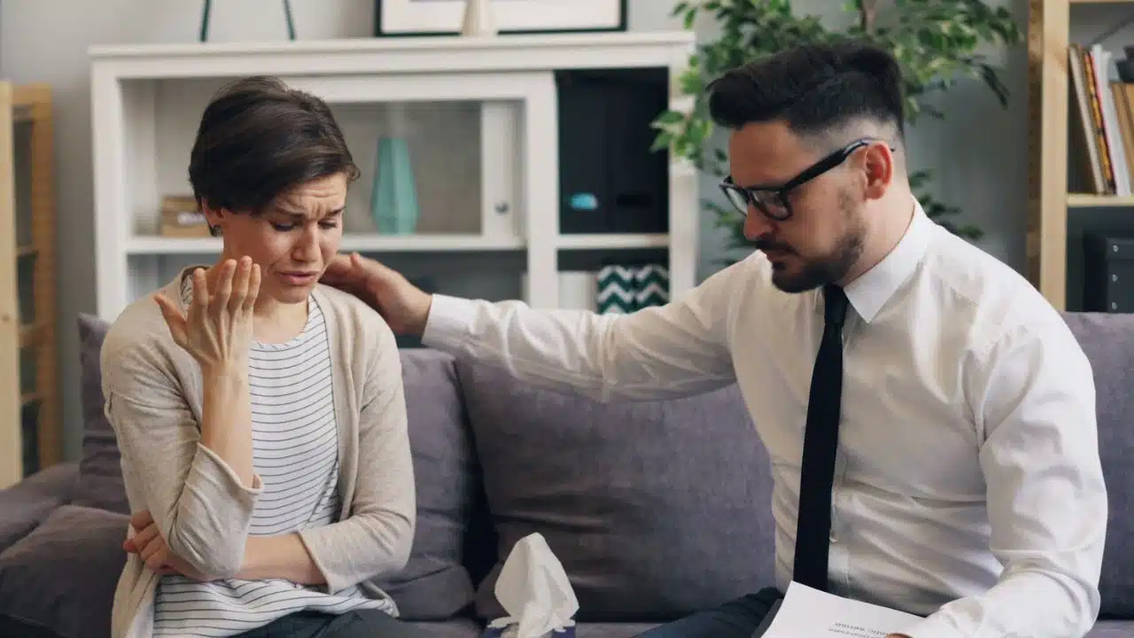 A man in a white shirt and tie comforts a crying woman on a sofa.