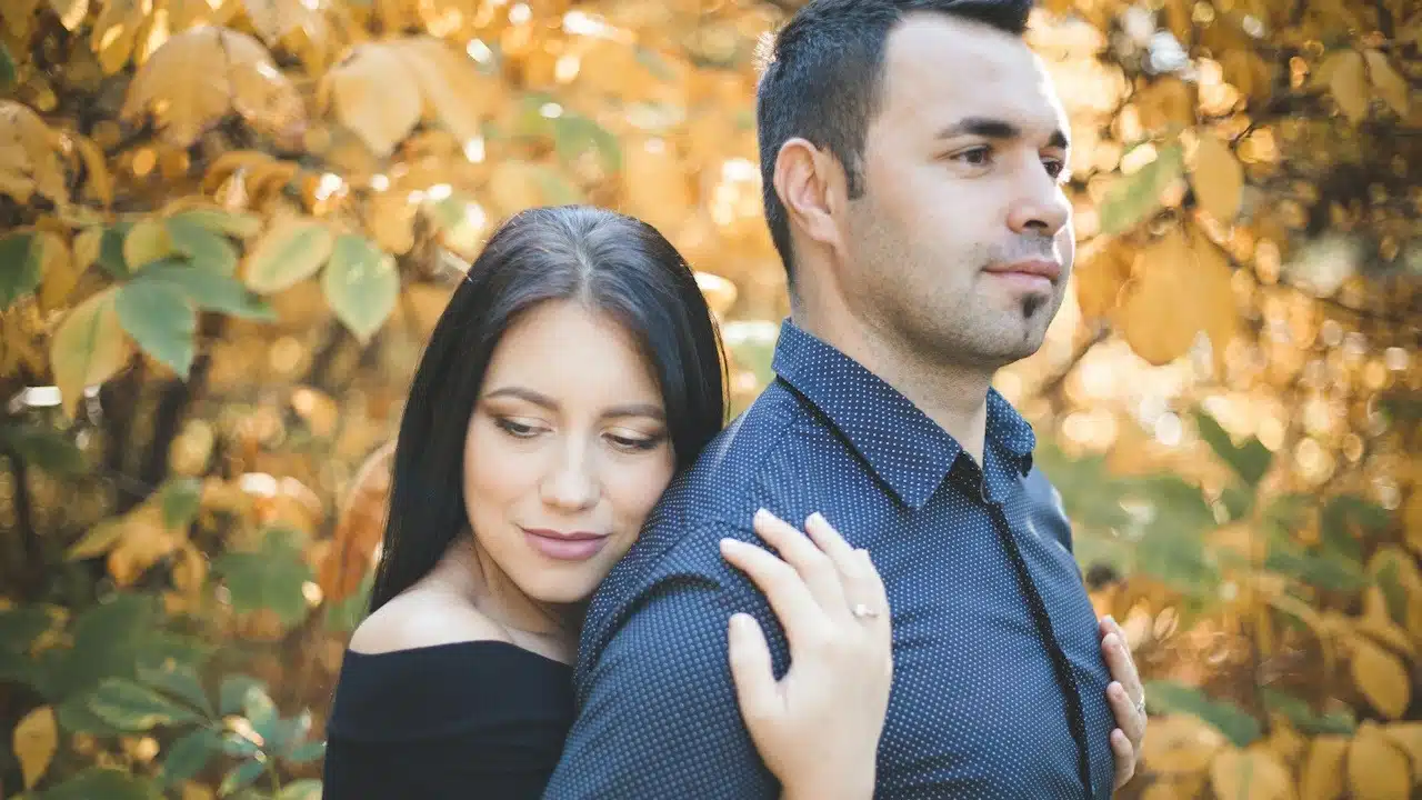 A woman hugging her husband from the backside while they are standing in autumn trees.