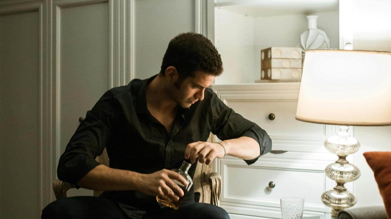 A man pouring a drink while sitting beside a lamp.