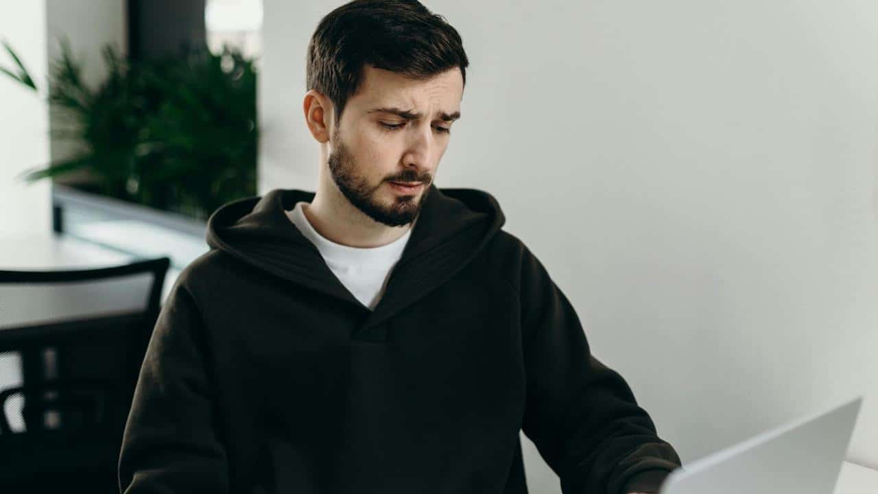 A man in a hoodie looking concerned while using a laptop.