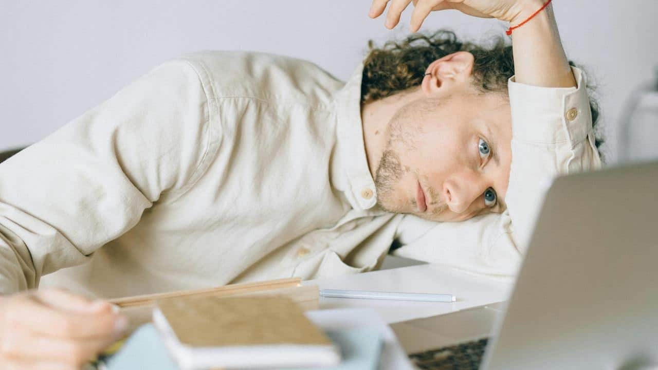 A tired man resting his head on his arm while looking at a laptop on a desk.