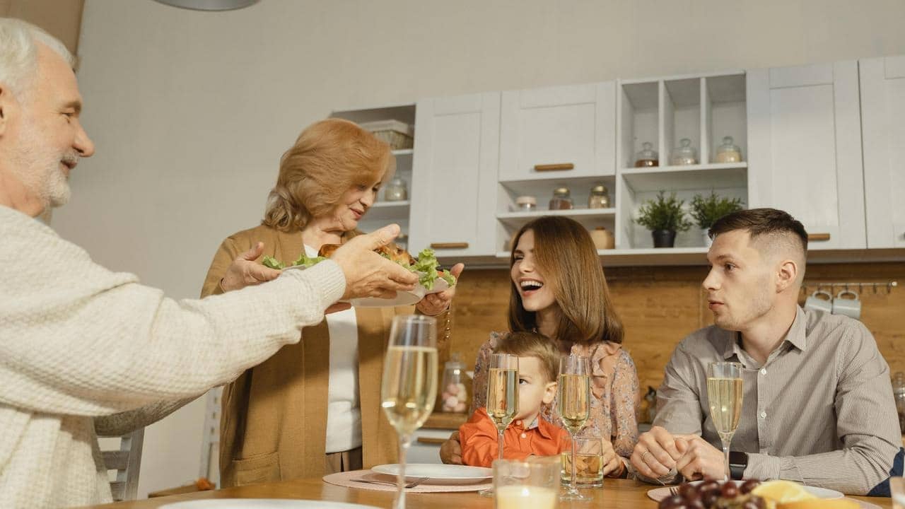 A family gathered around a dining table while an older woman serves a dish.