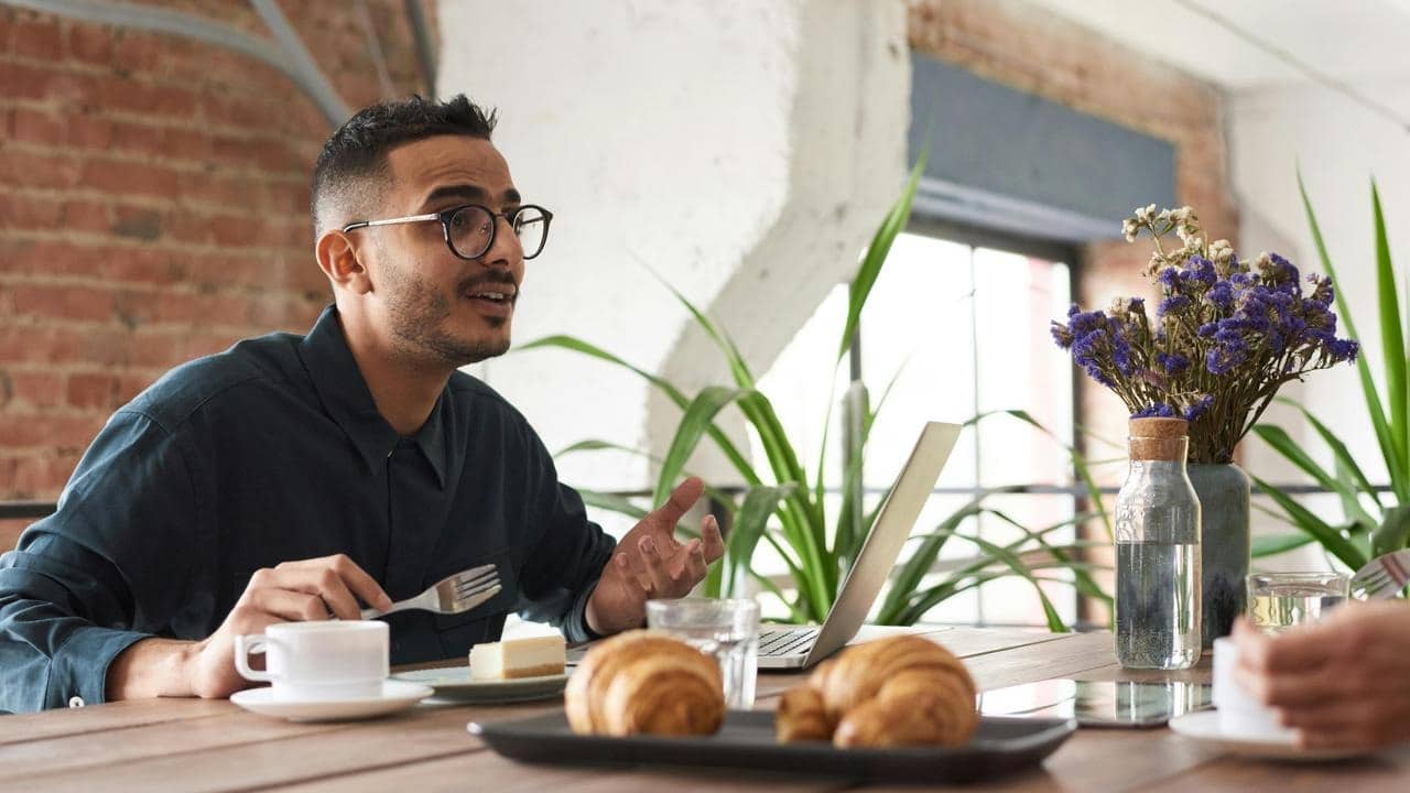 A man talking while sitting at a table with a laptop and breakfast items.