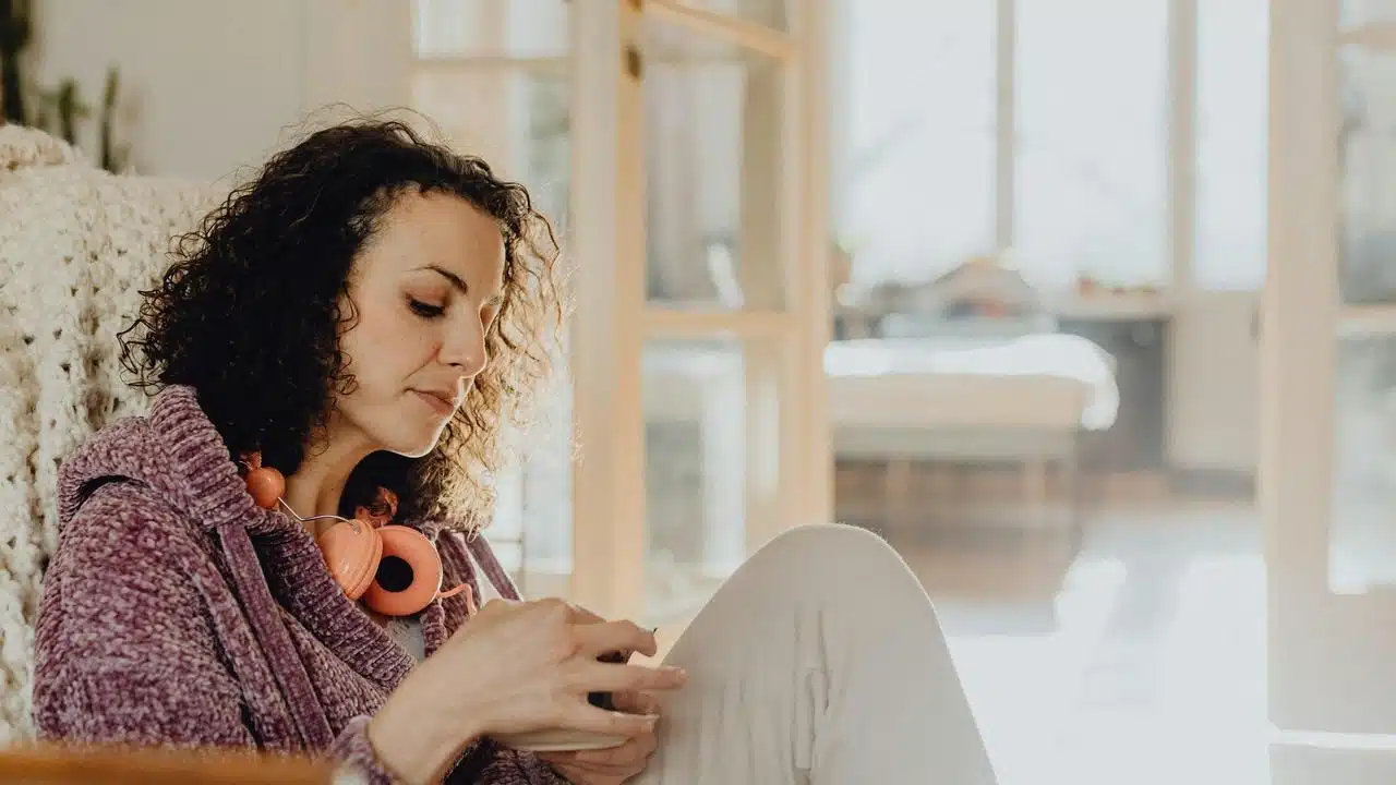 A woman sitting at home using her phone with headphones around her neck.