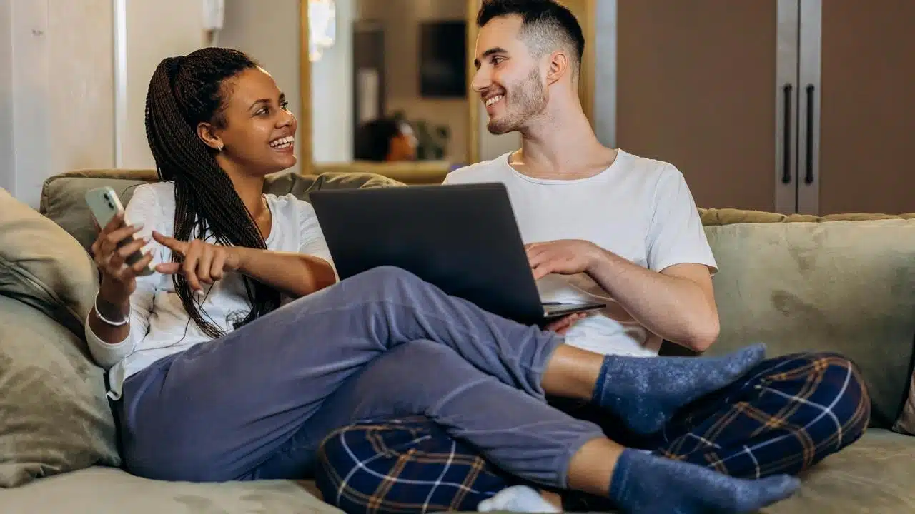 A couple sitting on a couch using a laptop and phone together.