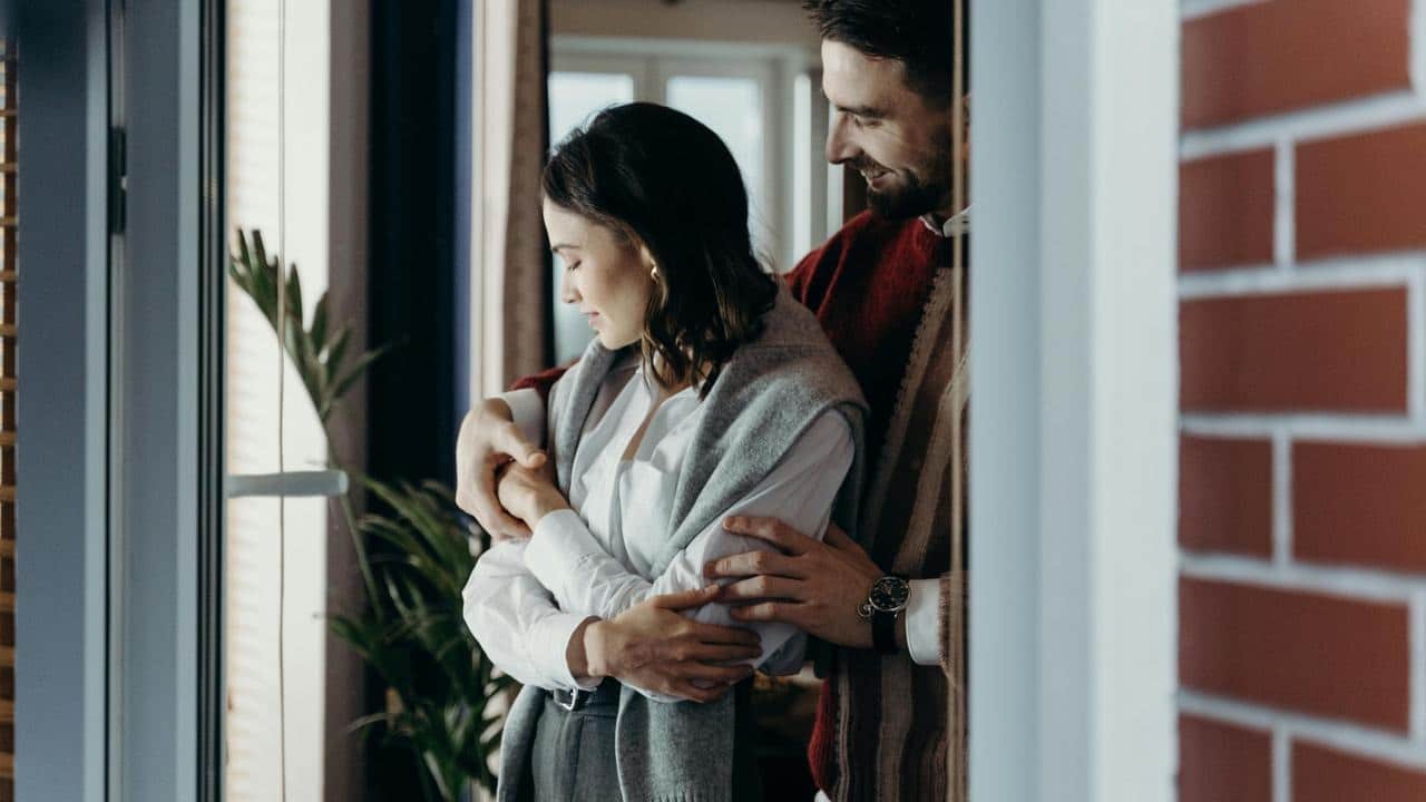 A couple standing by a window, gently embracing indoors.