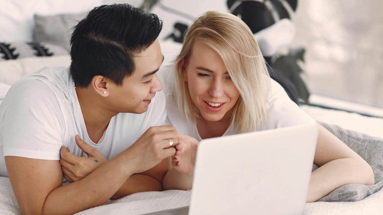 A couple lying on a bed and looking at a laptop together.
