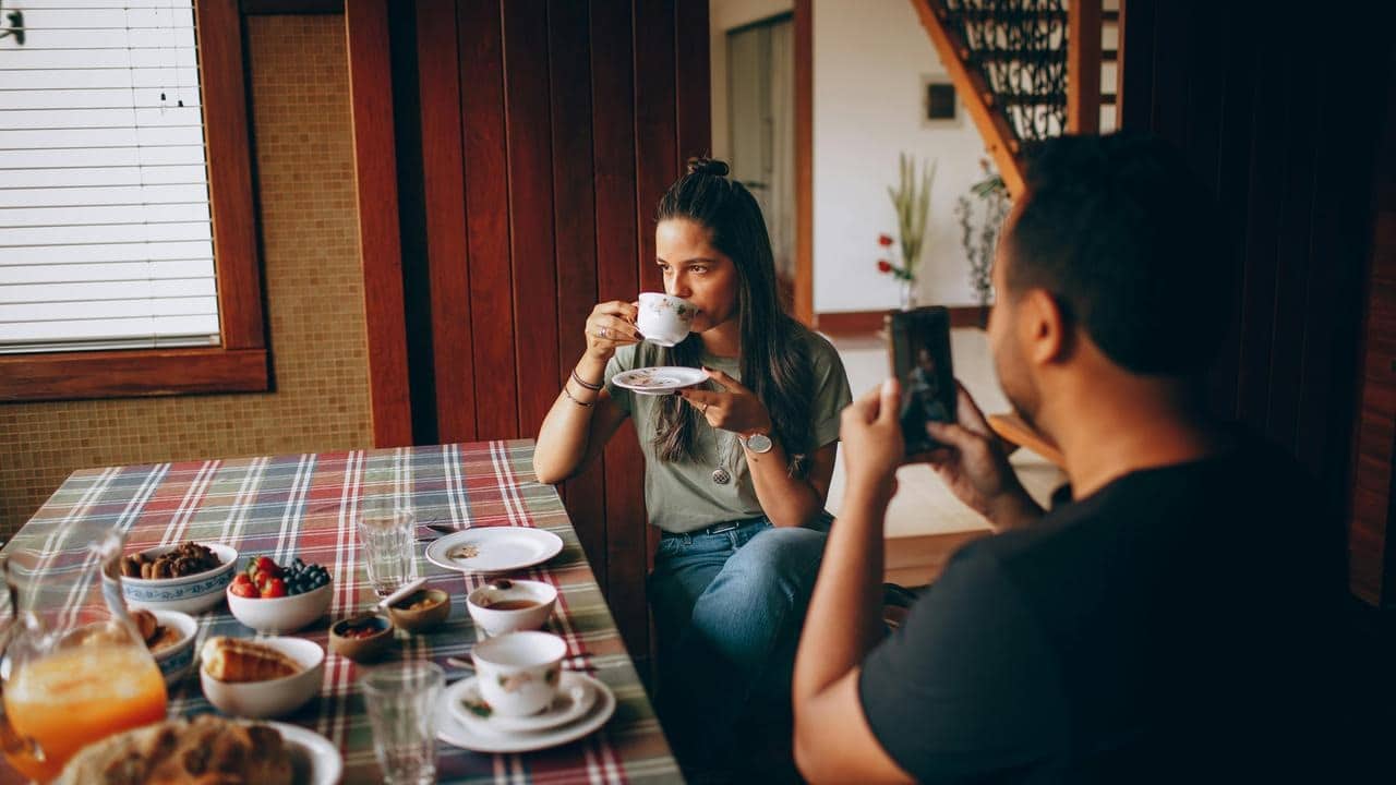 A woman drinking tea at a table while a man takes her photo.