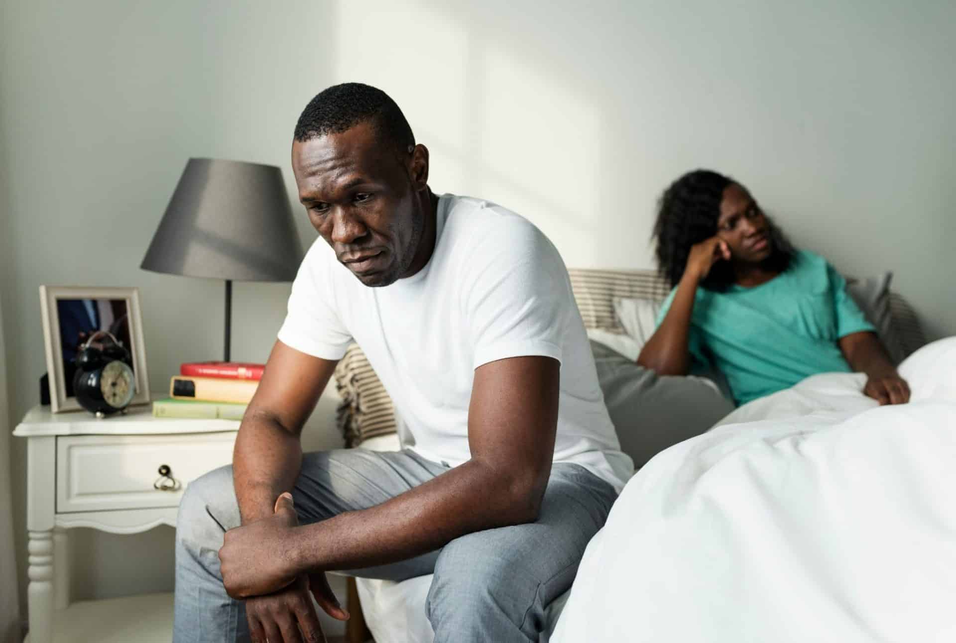 A man is sitting on the edge of a bed while a woman lies in bed behind him looking upset.