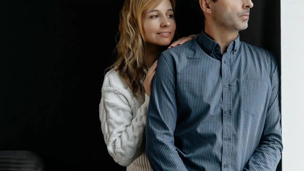 A woman gently holding a man’s shoulders as they stand by a window.