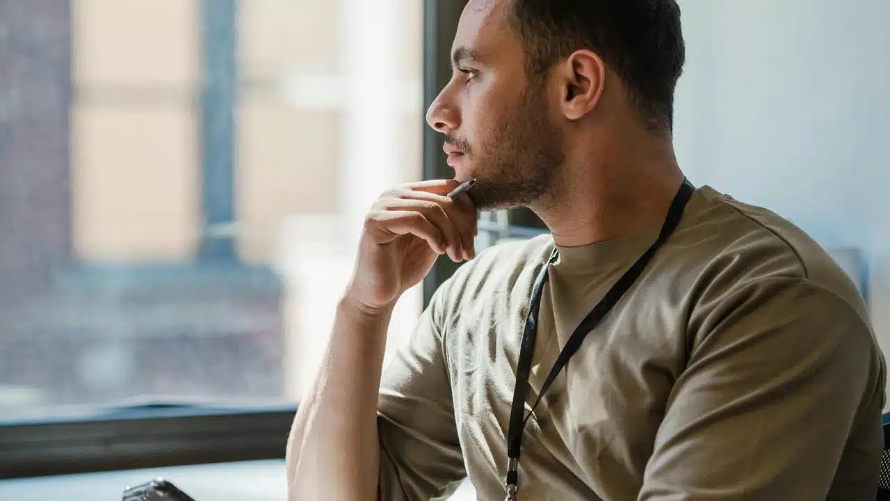 A man sitting by a window, holding a pen and looking thoughtful.