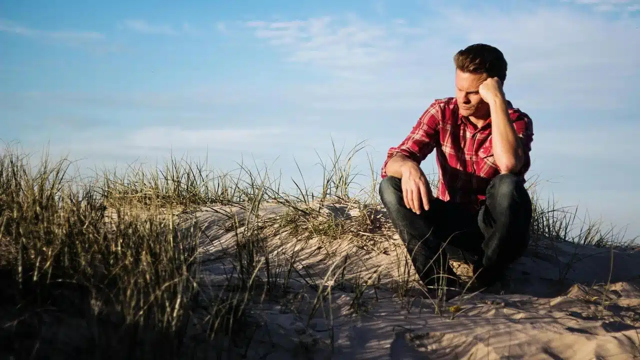 A man sitting on sandy dunes, looking thoughtful under a blue sky.