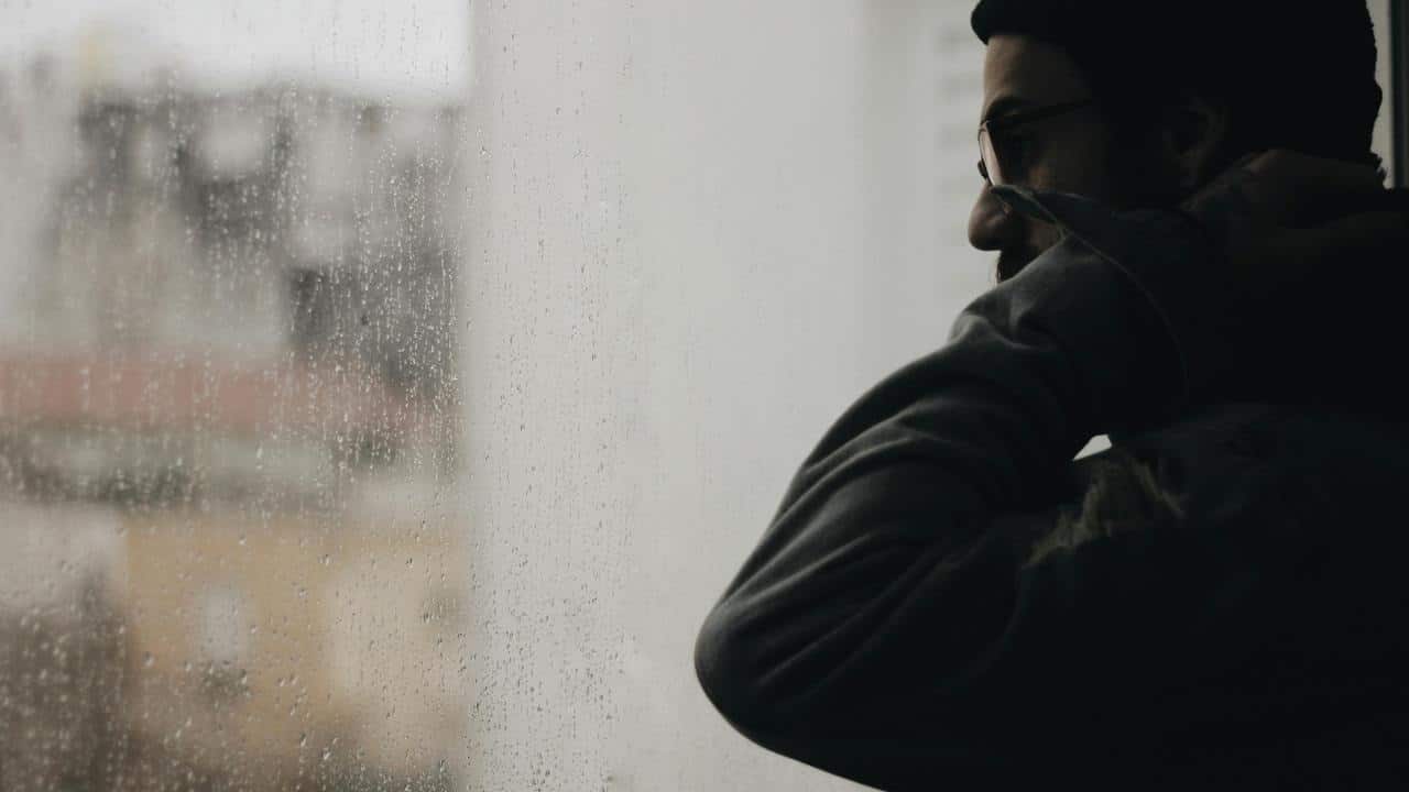 A man in a hoodie looking out a rain-covered window.